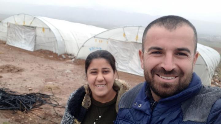  A man and a woman look into the camera. Behind them in a field are two large greenhouses with white tarpaulins. 