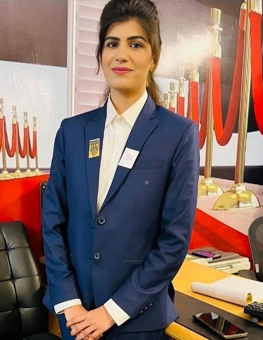  A woman in a blue uniform stands in front of a reception desk and smiles into the camera. 