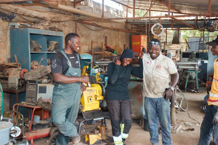  A young woman and several men are laughing together in a welding workshop.  